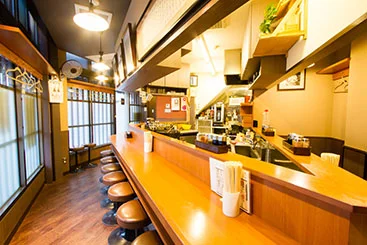 暖色の灯りがこぼれる島正の外観と、木のカウンターが奥へ続く店内を広角で写したアクセス写真